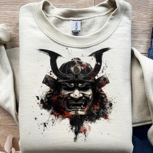 Puede incluir: Una sudadera crema con un gráfico detallado de una máscara de samurái. La máscara es predominantemente negra y blanca con detalles en rojo, mostrando detalles intrincados. La sudadera tiene cuello redondo y mangas largas, ideal para uso casual.