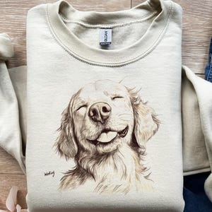 May include: A beige sweatshirt featuring a detailed illustration of a smiling golden retriever with its tongue out. The dog's eyes are closed, and the drawing is in shades of brown and beige. The word "Winking" is written in cursive.