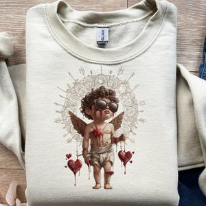 May include: A cream-colored sweatshirt featuring a graphic of a cherubic figure with sunglasses, wings, and blood-like streaks. The cherub holds two dripping hearts, set against a detailed circular design. The Gildan tag is visible at the neckline.