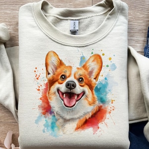 May include: A light beige sweatshirt featuring a watercolor illustration of a smiling corgi dog. The corgi has orange and white fur, with a pink tongue. Colorful paint splatters in red, blue, and orange surround the dog.