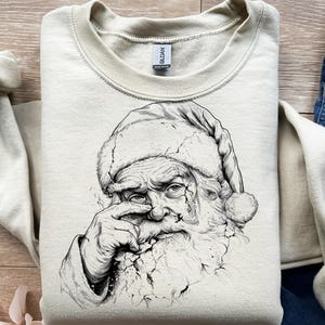 May include: A cream-colored sweatshirt featuring a detailed black and white illustration of Santa Claus. The graphic depicts Santa with a furrowed brow and a finger touching his nose, with a cracked effect.