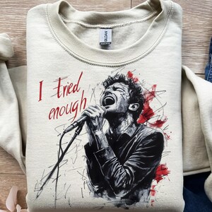 Può includere: Felpa beige con la scritta rossa "I tried enough". La grafica è un'illustrazione in bianco e nero di un uomo che canta in un microfono, con schizzi di vernice rossa.