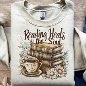 May include: Cream-colored sweatshirt with the text "Reading Heals the Soul." The design features a stack of books, a teacup, and daisies. The teacup has the words "tea's joy" on it.