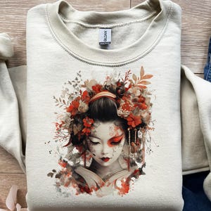 May include: A cream-colored sweatshirt featuring a watercolor illustration of a geisha with a floral headdress in shades of red, orange, and brown. The geisha has red eye makeup and red lips. The sweatshirt has a crew neck and long sleeves.
