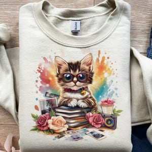 Puede incluir: Sudadera de color crema con un gráfico colorido de un gato con gafas, sentado sobre libros, con rosas y una cámara. El gato tiene pelaje marrón y blanco y lleva un collar rojo con una campana.