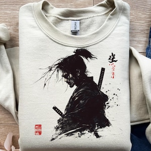 Puede incluir: Sudadera color crema con una ilustración en blanco y negro de un guerrero samurái con dos espadas. El diseño incluye texto japonés y un sello rojo. El guerrero está representado en una pose dinámica, con sombreado detallado y enfoque en la expresión del personaje.