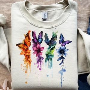 Puede incluir: Una sudadera de color crema con un diseño de acuarela de mariposas y flores. El diseño incluye mariposas y flores naranjas, moradas, verdes y azules. La sudadera tiene cuello redondo y mangas largas.