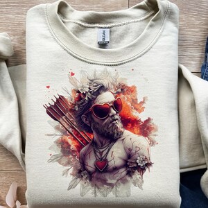Peut inclure: Sweat-shirt crème avec un graphisme d'un homme portant des lunettes de soleil en forme de cœur, une couronne de fleurs et un pendentif cœur. Le motif comprend des flèches et des accents rouges et oranges, parfait pour la Saint-Valentin.