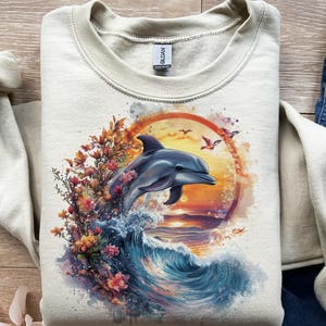 May include: A light beige sweatshirt featuring a vibrant graphic of a dolphin leaping from a wave, set against a sunset backdrop. The design includes colorful flowers and birds, creating a nature-themed aesthetic.