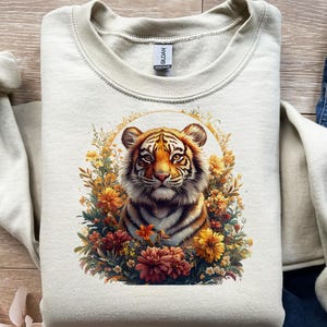 May include: Cream-colored sweatshirt featuring a detailed illustration of a tiger's face surrounded by colorful flowers. The tiger has orange and black stripes, with a focused gaze. The floral arrangement includes yellow, orange, and burgundy blooms.