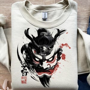 May include: A cream-colored sweatshirt featuring a black and red graphic of a Japanese demon mask. The design includes intricate details, with bold lines and splashes of red, and Japanese characters.