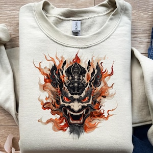 Könnte beinhalten: Ein cremefarbenes Sweatshirt mit einer detaillierten Grafik einer japanischen Dämonenmaske. Die Maske ist schwarz mit roten Augen und orangefarbenen Flammen. Das Sweatshirt hat einen Rundhalsausschnitt und ein "Gildan"-Etikett.