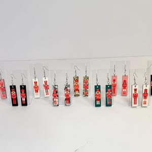 May include: A collection of rectangular dangle earrings featuring a toy soldier design. The earrings are in various colors, including pink, black, white, and green, with silver-tone ear wires. The toy soldier design is in red, black, and white.