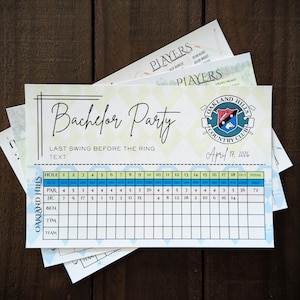 May include: A pile of golf scorecards with the text "Bachelor Party" and "Last Swing Before The Ring". The cards display a score table, the Oakland Hills Country Club logo, and the date April 17, 2026. The background is a dark wood surface.