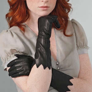 May include: A pair of black leather gloves extending past the wrist, featuring a distinct zigzag or serrated edge design at the cuff. The gloves are constructed with visible stitching along the fingers and a smooth, matte finish on the leather surface.