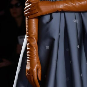 Peut inclure: Une robe bleu ardoise à manches courtes et jupe plissée, ornée d'un motif répétitif de petits cercles blancs. La robe est associée à de longs gants d'opéra en cuir fauve, lisses, qui dépassent les coudes, créant un contraste de texture.