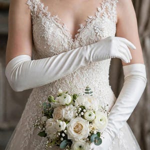 May include: An ivory lace gown with a V-neckline and intricate floral embroidery featuring pearl accents, paired with long, smooth satin opera gloves and a bouquet of white roses, ranunculus, and small green eucalyptus leaves.