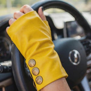May include: A bright yellow fingerless leather glove featuring three round, metallic bronze-toned buttons aligned vertically along the wrist area. The glove has a smooth texture and covers the hand while leaving the fingers exposed.