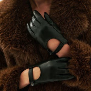 Könnte beinhalten: Schwarze Lederhandschuhe mit fingerlosem Design und Riemendetail. Die Handschuhe sind vor einem Hintergrund aus braunem Fell platziert, was einen Kontrast in Texturen und Farben erzeugt. Die Handschuhe scheinen neu zu sein.