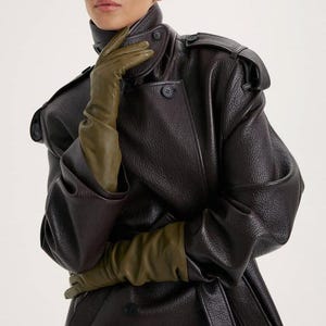 Peut inclure: Un trench-coat en cuir texturé marron foncé doté d'un col haut, de pattes d'épaule et de boutons sombres, associé à de longs gants en cuir vert olive qui dépassent les poignets, créant un effet superposé avec les manches larges du manteau.