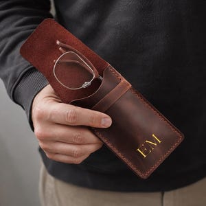 May include: A dark brown leather glasses case with a flap closure and visible stitching along the edges. The front of the case features the gold embossed initials EM. A pair of thin-framed eyeglasses is partially inserted into the leather sleeve.