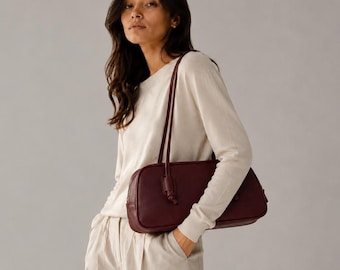 May include: A dark burgundy rectangular shoulder bag with a smooth finish and two thin, long straps. The bag features a structured, rounded silhouette with a top zipper closure and is carried under the arm against a cream-colored long-sleeve top.