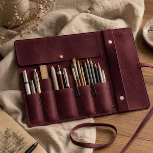 May include: A deep burgundy leather roll-up case featuring multiple interior slots filled with various writing instruments, including pens, pencils, and a small wooden ruler. The case is secured with a flap and a long leather strap for closure.