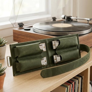 Op de afbeelding: Een donker olijfgroene lederen mondharmonikahoes met vier afzonderlijke vakken, elk met een zilverkleurige metalen mondharmonika. De hoes is voorzien van een verstelbare lederen riem met een metalen gesp om de instrumenten veilig te houden.