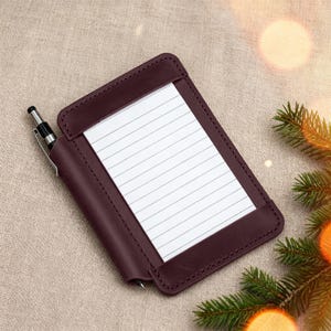 May include: A burgundy leather notepad holder with a pen. The notepad has lined paper and is held in place by a leather frame. The pen is silver and black. The notepad holder is on a neutral-coloured surface.
