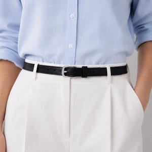 May include: A slim black leather belt with a silver-toned metal buckle, worn over white trousers and a light blue button-down shirt. The belt features a simple prong closure and a single matching leather keeper loop to secure the strap end.