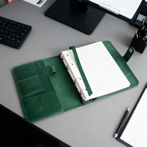 May include: Open green leather binder with white paper pages. The binder is open, revealing the interior pockets and a green ribbon bookmark. The binder is on a gray desk with a keyboard and monitor in the background.