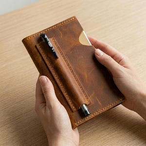Puede incluir: Una funda rectangular para cuaderno de cuero marrón con un bolsillo vertical cosido en la parte delantera. Un bolígrafo de gel negro está insertado en el bolsillo, con la punta visible en la parte inferior y el clip en la parte superior.