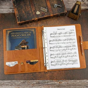 May include: A tan leather music binder featuring a three-ring mechanism holding sheet music for Clair de Lune, a pocket containing an Advanced Classical Piano Solos book, two small cards, and a gold-toned pen attached to the side.