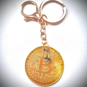 Può includere: Portachiavi Bitcoin color oro con un ciondolo a forma di moneta rotonda. La moneta presenta il simbolo Bitcoin e testo attorno al bordo. Il portachiavi include un moschettone, una catena e un portachiavi.