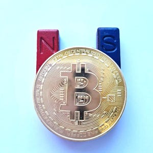 Può includere: Una moneta Bitcoin d'oro è posizionata tra un magnete rosso 'N' e un magnete blu 'S'. La moneta presenta il simbolo Bitcoin e testo sul bordo, tra cui 'BITCOIN-DIGITAL-DECENTRALIZED-PEER'. Lo sfondo è azzurro.