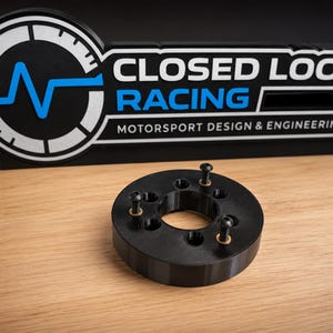 May include: A black circular steering wheel adapter with three small metal screws inserted into the top surface. Behind it stands a black sign with white and blue text that reads CLOSED LOOP RACING MOTORSPORT DESIGN & ENGINEERING.