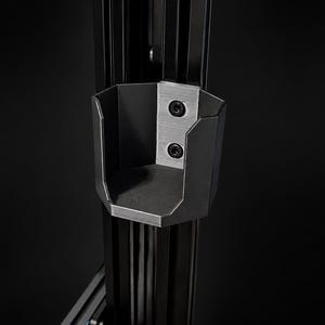 May include: A black metal bracket with a hexagonal shape, attached to a vertical support structure. The bracket has a small, open compartment and is secured with two black screws. The background is a solid black.