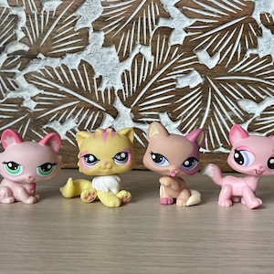 May include: Four colorful Littlest Pet Shop cat figurines. The cats are in various poses and colors, including pink, tan, and yellow. Each cat has large eyes and unique markings. The background features a carved wooden panel.
