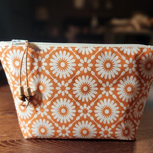 May include: Cosmetic bag with an orange and white floral pattern and a zip closure. The bag has a rectangular shape with a beige zip and a drawstring with wooden beads. It is on a wooden surface.