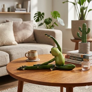 May include: A green ceramic frog figurine in a yoga pose, with arms extended upwards and legs spread. The frog has gold accents on its hands and feet. It sits on a round wooden coffee table, with books and a cactus nearby.