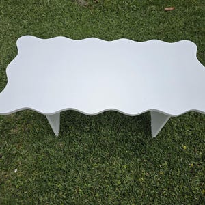 May include: A white, scalloped-edge coffee table with two triangular legs. The table is made of wood and sits on a bed of green grass. The table's unique shape and design make it a stylish addition to any living space.