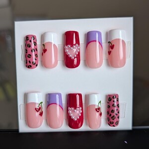 May include: A set of ten artificial nails in various designs. The nails feature a mix of pink, red, and purple colors, with accents like a leopard print, cherry designs, and pearl heart embellishments. The nails are displayed on a white card.