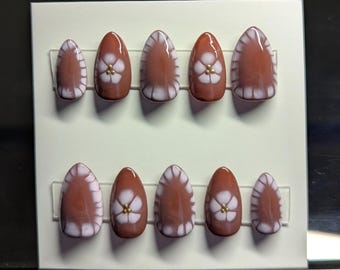 May include: A set of almond-shaped, press-on nails. Each nail is a warm brown color with a white, scalloped design along the edges. A white flower with a gold center is painted on each nail. The nails are arranged on a white card.