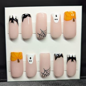 May include: A set of ten press-on nails with a light pink base. Each nail features Halloween-themed designs, including bats, spiderwebs, ghosts, and pumpkins. The nail art is in black, white, and orange.