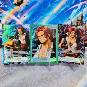May include: Three One Piece trading cards featuring the character Shanks. Each card displays the name Shanks, a power level of 5000, and a life value of 5. The cards showcase detailed anime-style illustrations of the red-haired character in various poses.