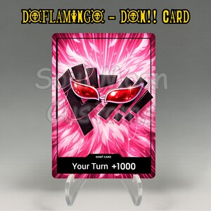 May include: A rectangular card with the text "DOFLAMINGO - DON!! CARD" at the top. The card features a pink feathery background with a black and red graphic. The bottom of the card reads "Your Turn +1000".