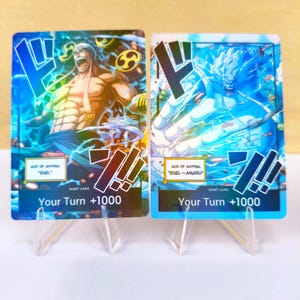 Puede incluir: Dos cartas coleccionables con ilustraciones de personajes y efectos de energía. Las cartas muestran texto como "GOD OF SKYPIEA", "ENEL" y "Your Turn +1000". Una carta tiene un diseño amarillo y marrón, la otra es predominantemente azul.