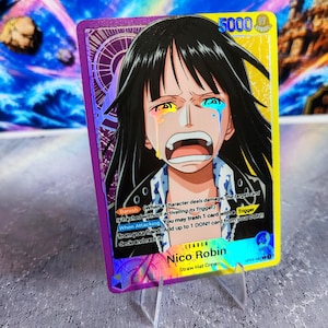 K&ouml;nnte beinhalten: Eine One Piece Sammelkarte mit Nico Robin mit schwarzen Haaren und einem weinenden Gesichtsausdruck. Die Karte hat einen violetten und gelben Rand, eine St&auml;rke von 5000 und Text, der die F&auml;higkeiten Banish und When Attacking beschreibt.