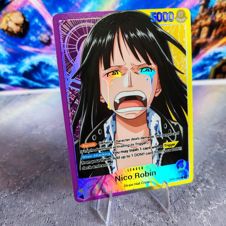 K&ouml;nnte beinhalten: Eine One Piece Sammelkarte mit Nico Robin mit schwarzen Haaren und einem weinenden Gesichtsausdruck. Die Karte hat einen violetten und gelben Rand, eine St&auml;rke von 5000 und Text, der die F&auml;higkeiten Banish und When Attacking beschreibt.