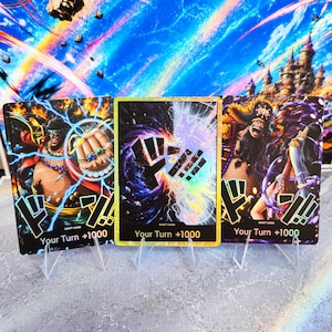 Pode incluir: Três cartas colecionáveis retangulares com ilustrações de personagens de One Piece e uma carta Don! central. Cada carta exibe o texto Your Turn +1000 na parte inferior. As cartas apresentam ilustrações vibrantes e detalhadas com fundos escuros e energéticos.